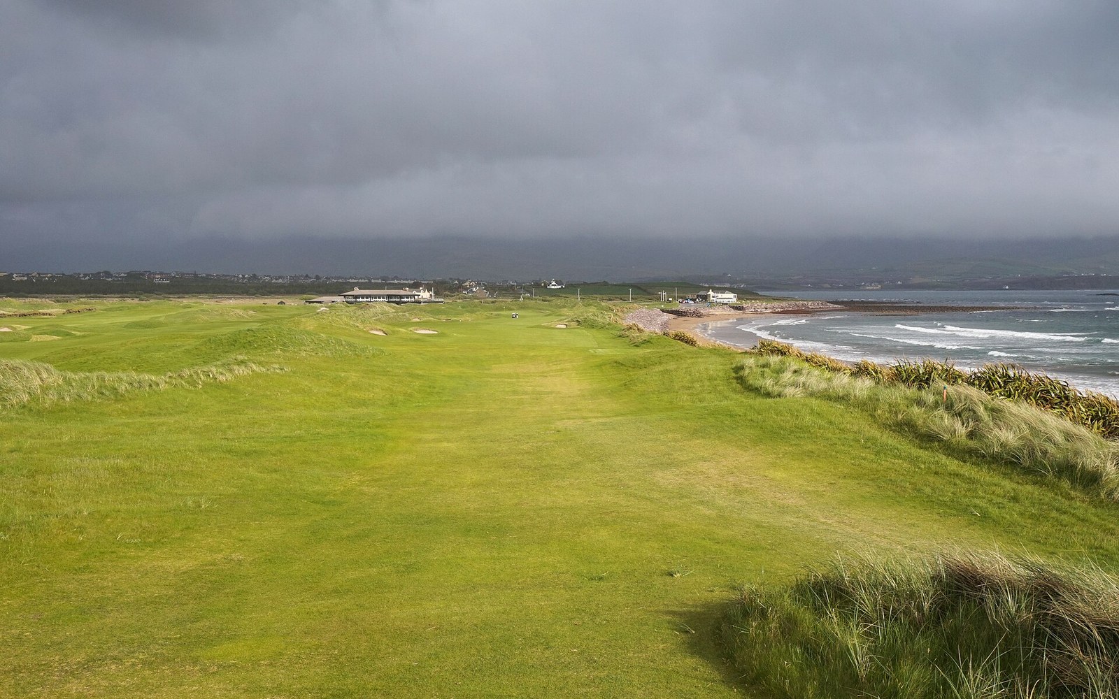 A coastal links fairway runs alongside the ocean under a stormy grey sky